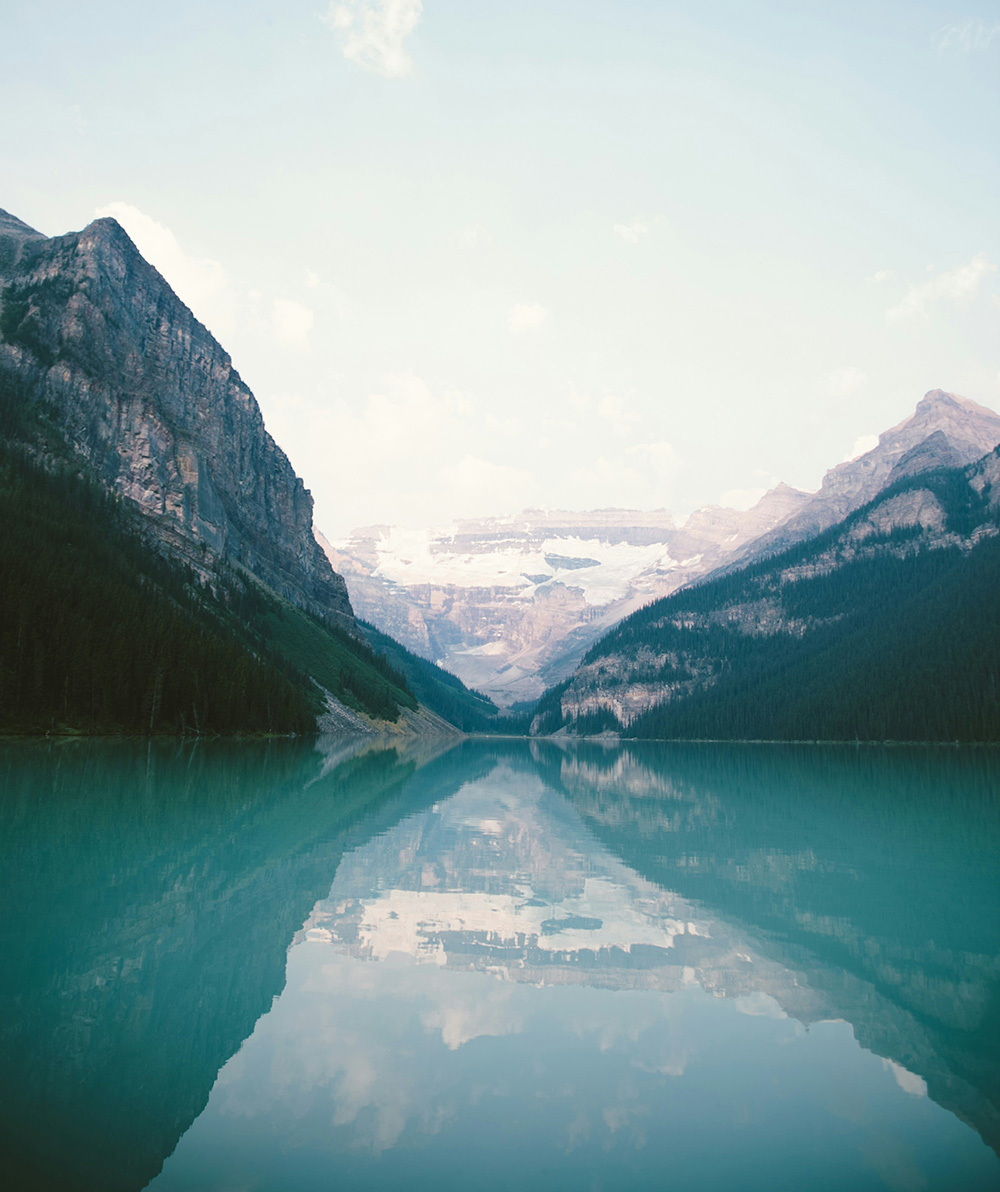 A serene mountain lake with turquoise water reflecting the surrounding forested peaks and a partly cloudy sky.