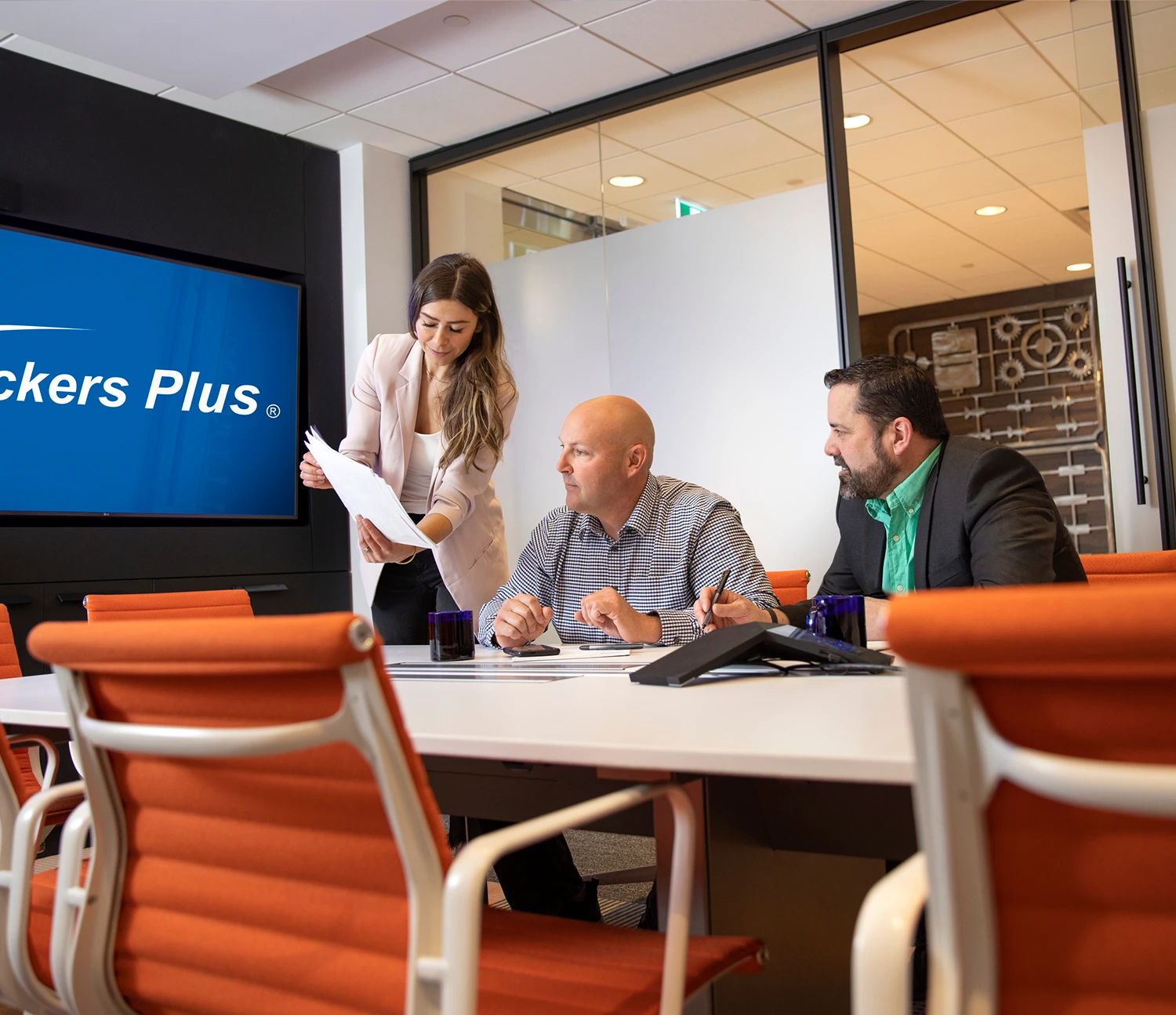 Three people sit at a conference table in a modern office, engaged in discussion. A television on the wall displays the Packers Plus logo.